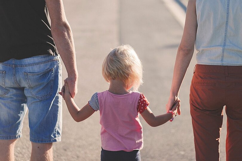 Ein Vater, ein Kind und eine Mutter gehen Hand in Hand. Man sieht sie von hinten. Der Vater träge eine knielange Jeans und ein blaues T-Shirt, das Kind ein rosa T-Shirt und die Mutter eine rote Jeans und ein weißes T-Shirt. 