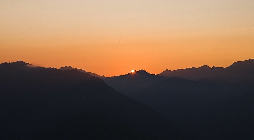 Abenstimmung in den Tiroler Bergen: Dunkle Bergketten, oranger Himmel und die Sonne als kleiner, gelber Punkt in der Mitte des Bildes und am Rand der Berge.