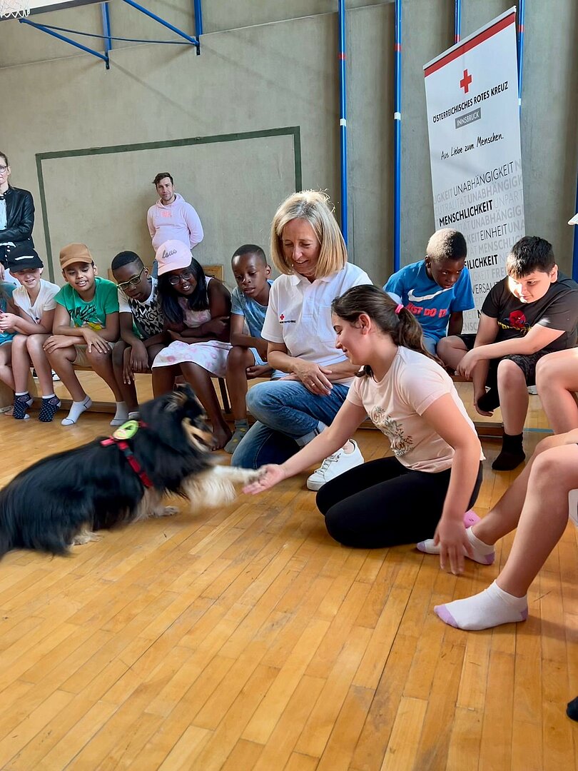 Im Turnsaal der Volkschule Neuarzl: Viele Kinder sitzen auf den Langbänken. Caspar gibt einem Kind seine rechte  Pfote. Cornelia Walder sitzt beobachtend daneben. 