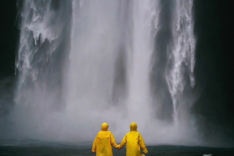 Zwei Menschen stehen mit knallgelben Regenmänteln händehaltend vor einem tosenden Wasserfall.
