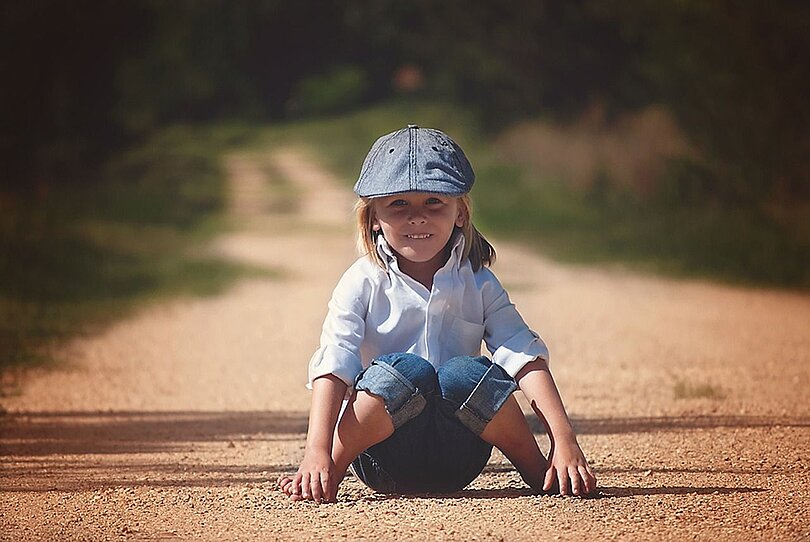 Ein Junge sitzt barfuss auf einem Feldweg. Er trägt eine Jean, ein weißes Hemd und eine blaue Mütze und ein Lächeln im Gesicht, das zeigt, dass es ihm gut geht.
