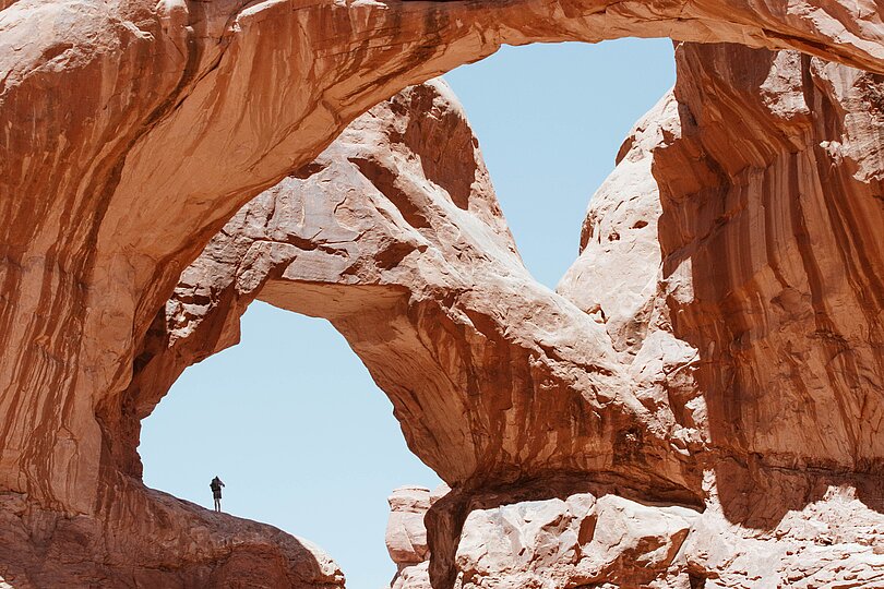 Doppelbogen in rotem Gestein eines amerikanischen Nationalparks. Ein Mensch steht in einem Bogen vor blauem Hintergrund.