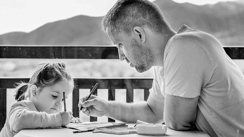 Wahrscheinlich sitzen ein Vater und seine Tochter gemeinsam am Tisch und zeichnen miteinander oder schreiben etwas gemeinsam.