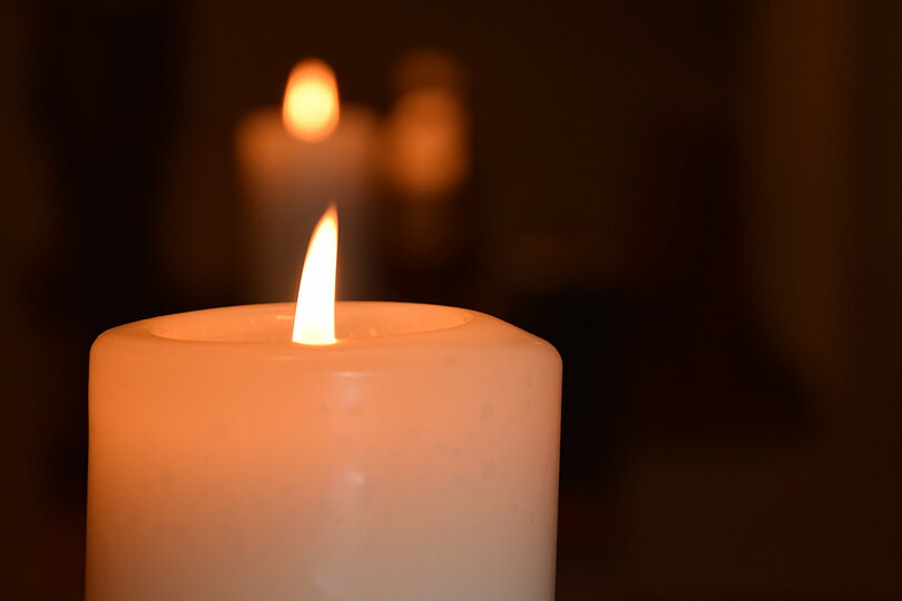 Das Licht einer großen, weißen Kerze. Im Hintergrund sieht man noch eine Kerze brennen. 