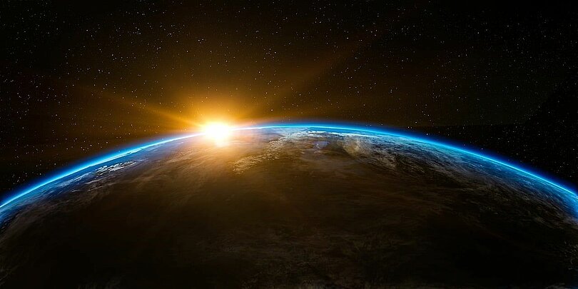 Die Erde vom Weltall aus gesehen. Man sieht den oberen Teil der Erdkugel. Im Hintergrund blinzeln aus dem Dunkeln die ersten Sonnenstrahlen über die Erdkugel. 