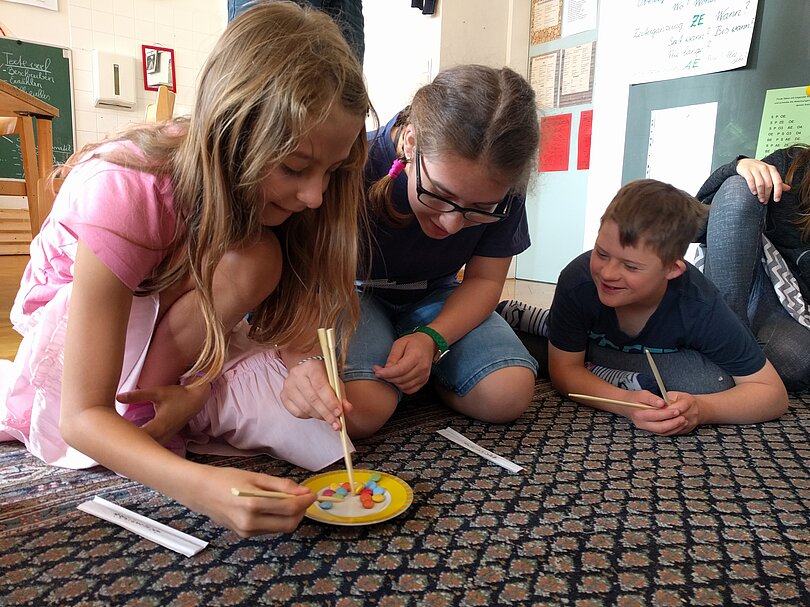 Drei Kinder sitzen und liegen am Boden. Vor ihnen steht ein Teller mit Smarties. Mit Stäbchen greifen sie die Smarties. 