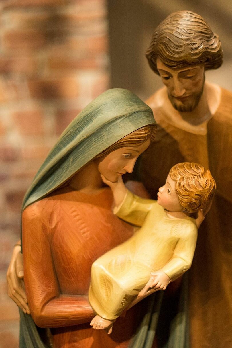 Die heilige Familie als geschnitzte Figuren. Maria hält das Kind in ihren Händen und schaut es liebevoll an Josef steht hinter den beiden und legt seinen Arm schützend und bestärkend auf Maria. 