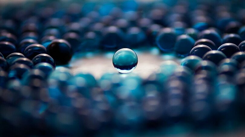 Am Tisch liegen blaue Glasmurmeln. In der Mitte ist ein Kreis frei und ganz in der Mitte liegt eine blaue Murmel allein.