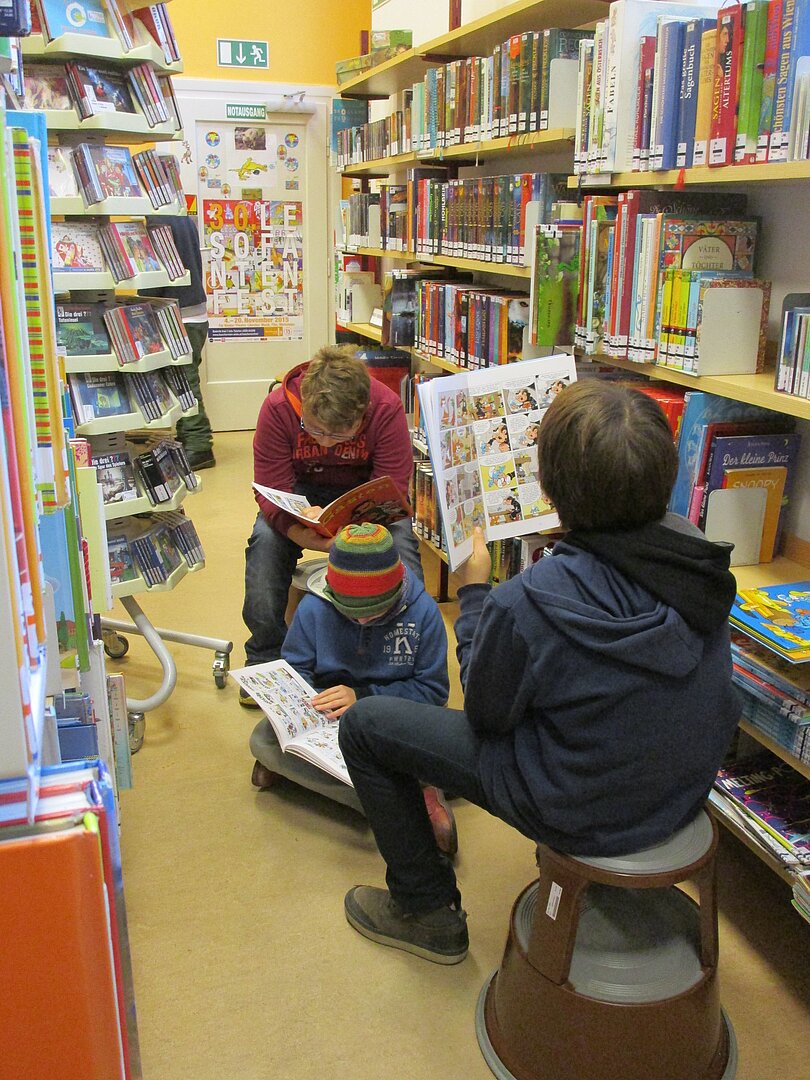 3 Kinder sitzen lesend in einem schmalen Gang der Bibliothek. 