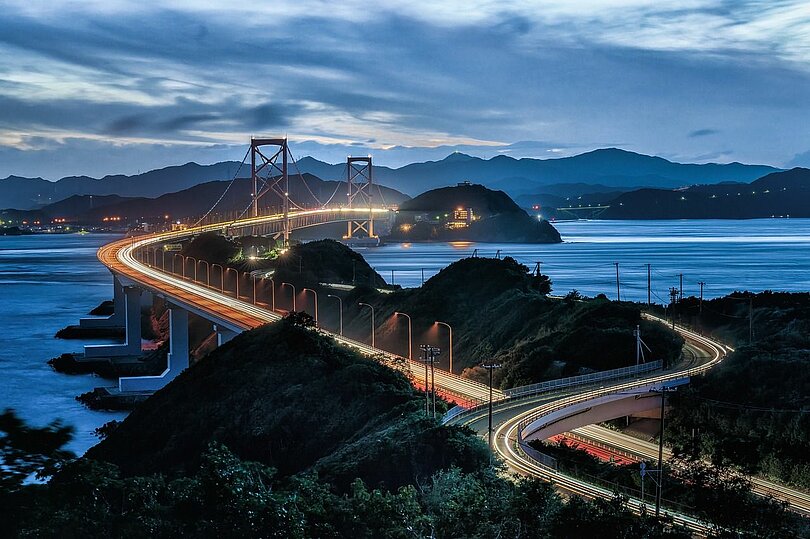 Eine Brücke spannt sich über eine Bucht. Eine zweite Straße zieht sich teilweise parallel. Abendstimmung und die man sieht von den fahrenden Autos nur die langen Lichter. 