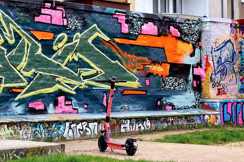 Street Art: Graffity auf einer Mauer mit orange, pink, blau, grün und gelb. Ein rosa E-Scooter steht davor. 