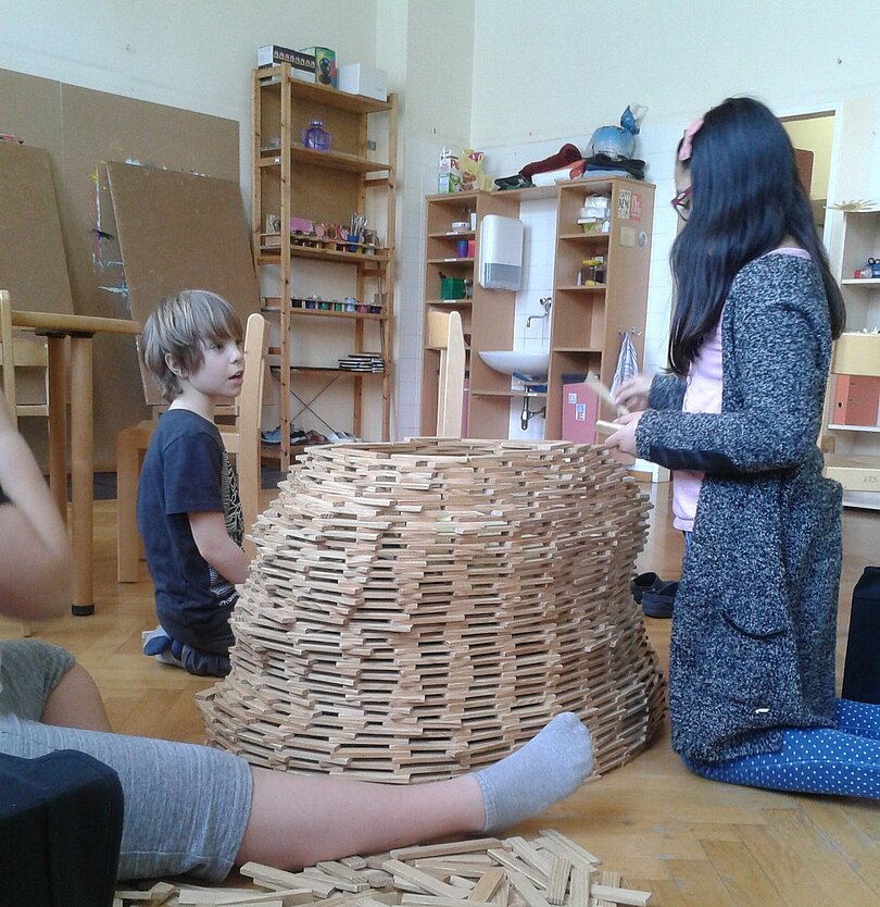 In der Mitte steht ein Kapplaturm. Zwei oder wahrscheinlich drei Kinder unterschiedlichen Alters sitzen rund herum und sind ganz konzentriert beim Bauen. 