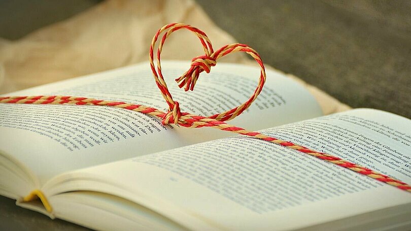 Ein Buch liegt offen auf einem Tisch. Rund um das Buch ist eine gelb-rote Schnur. In der Mitte des Buches ist die Schnur zu einem Herz geformt. 