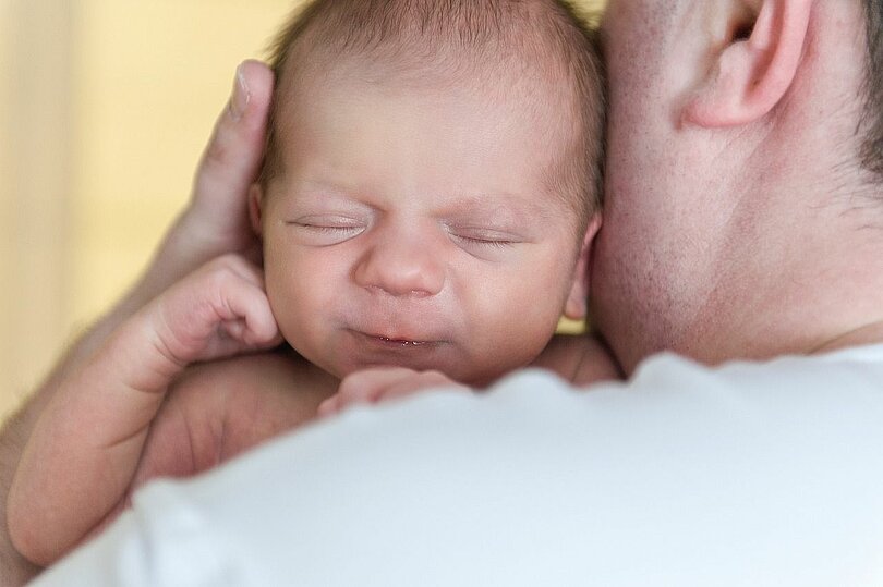 Ein Vater hält sein neugeborenes Kind. Der Kopf des Kindes liegt ganz zärtlich an der Wange des Vaters. Mit der linken Hand stützt der Vater behutsam den Kopf des Kindes. Das Baby hat die Augen geschlossen und zufrieden.