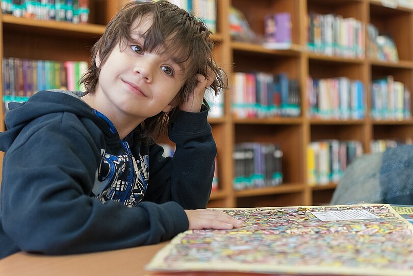 Bub sitzt am Tisch und lächelt verschmitzt. Er hat wohl ein Puzzle fertig gestellt. 