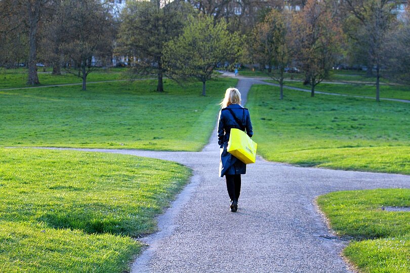 Eien Frau in dunklem Mantel und mit gelber Tasche geht in einem Park auf eine Kreuzug zu. 