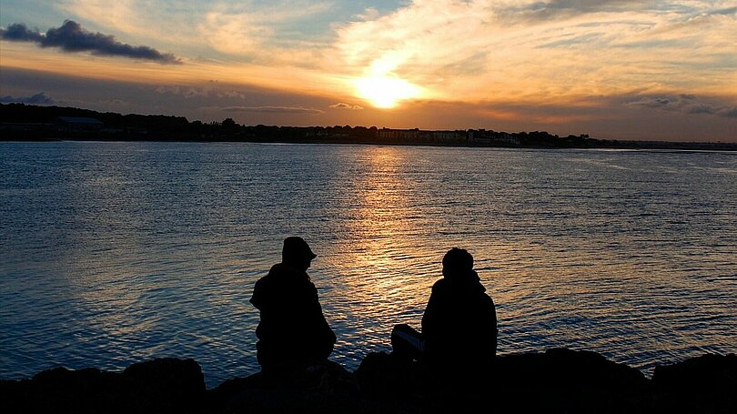 Zwei Menschen sitzen am Rand eines Sees und schauen in Richtung untergehende Sonne. Man hat den Eindruck, dass sich die beiden unterhalten. Zu sehen sind sie im Gegenlicht als schwarze Umrisse. 