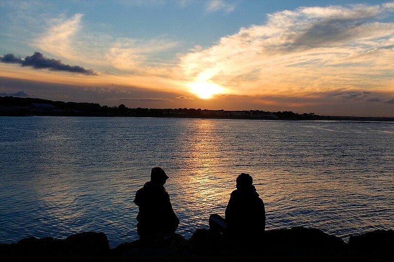 Zwei Menschen sitzen am Rand eines Sees und schauen in Richtung untergehende Sonne. Man hat den Eindruck, dass sich die beiden unterhalten. Zu sehen sind sie im Gegenlicht als schwarze Umrisse. 