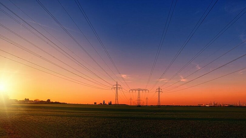 Stromleitungen führen zu drei Strommasten hin, die in einiger Entfernung stehen. Im Bild links geht die Sonne. Die Farben des Bildes: die Wiese wirkt graugrün. Der Horizont rot und der Himmel dunkelblau. 