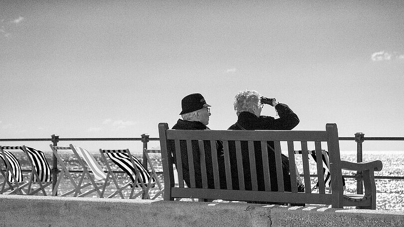 Ein älteres Paar sitzt auf einer Bank und schaut aufs Meer. Es ist sonnig, aber windig. Links neben der Bank stehen ein paar wahrscheinlich rot-weiß gestreifte Liegestühle. 
