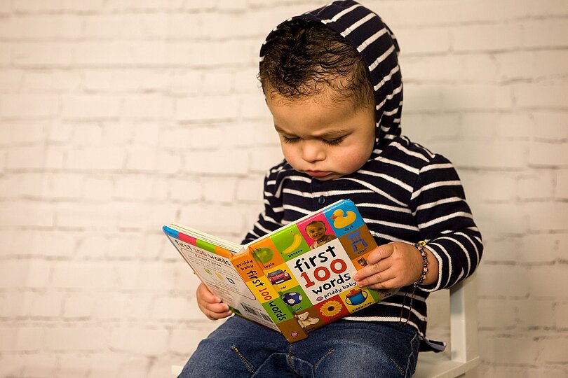 Ein Kind (ca. 2 Jahre) sitzt auf einem weißen Stuhl und liest ganz konzentriert ein Bilderbuch