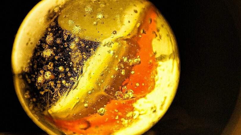 Ein weiße Glasmurmel mit einem orangen Streifen wird von hinten her angeleuchtet und leuchtet golden.