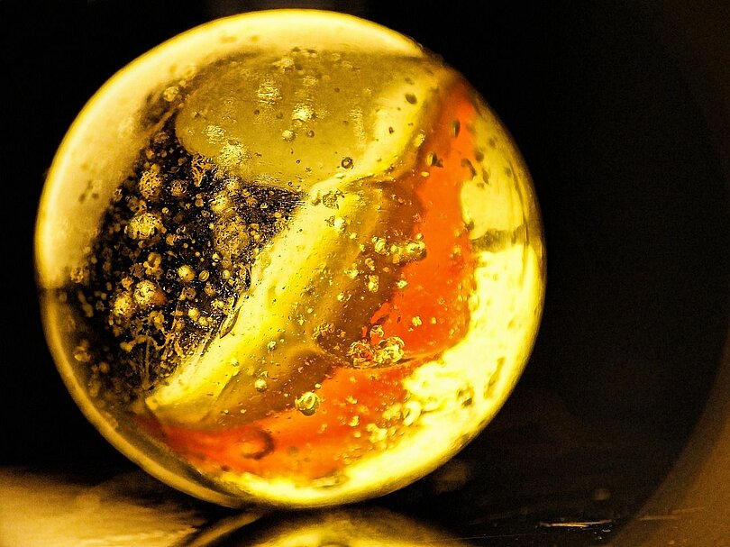 Ein weiße Glasmurmel mit einem orangen Streifen wird von hinten her angeleuchtet und leuchtet golden.