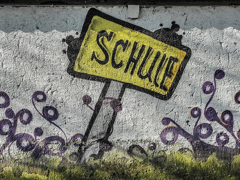 Streetart: Ein Holzpflock, ein gelbes Schild auf dem in Großbuchstaben SCHULE steht.