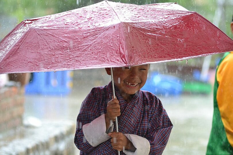 Ein asiatisch aussehender Junge in lila Gewand mit weißen Ärmeln steht lachend unter einem rosa Regenschirm, während es regnet. 