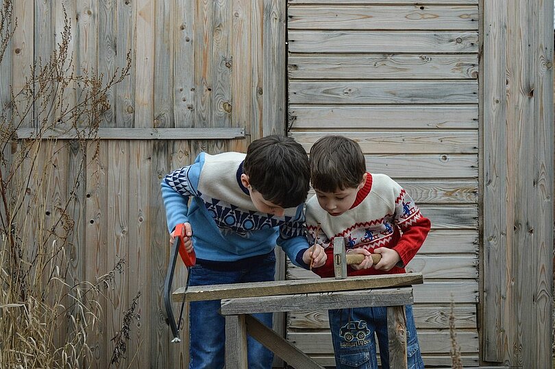 Zwei Buben mit bunten Pullovern stehen vor einer grauen Holzwand und suchen einen geeigneten Platz um einen Nagel in zwei Bretter, die auf Schragen liegen,  einzuschlagen