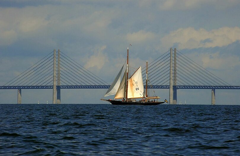 Ein Segelschiff fährt im blauen Meer vor der Öresundbrücke. Am blauen Himmel ziehen leichte weiße Wolken. 