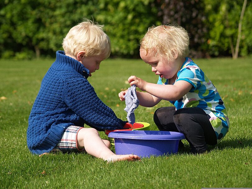 Zwei Kinder spielen auf einer Wiese mit zwei Plastikwannen und Waschlappen 