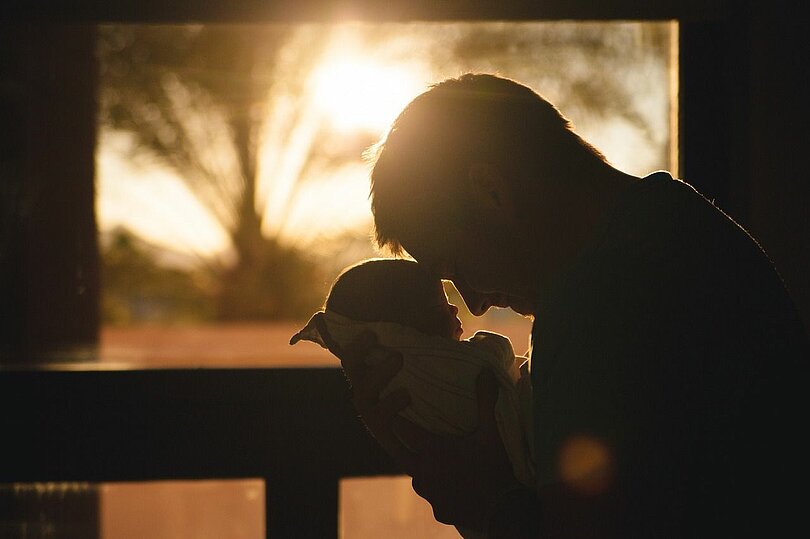 Ein Vater schaut sein neugeborenes Kind an und berührt mit seiner Stirn, die Stirn des Kindes. Im Hintergrund geht die Sonne hinter Palmen unter. Von den Figuren im Vordergrund sieht man die dunklen Umrisse. 